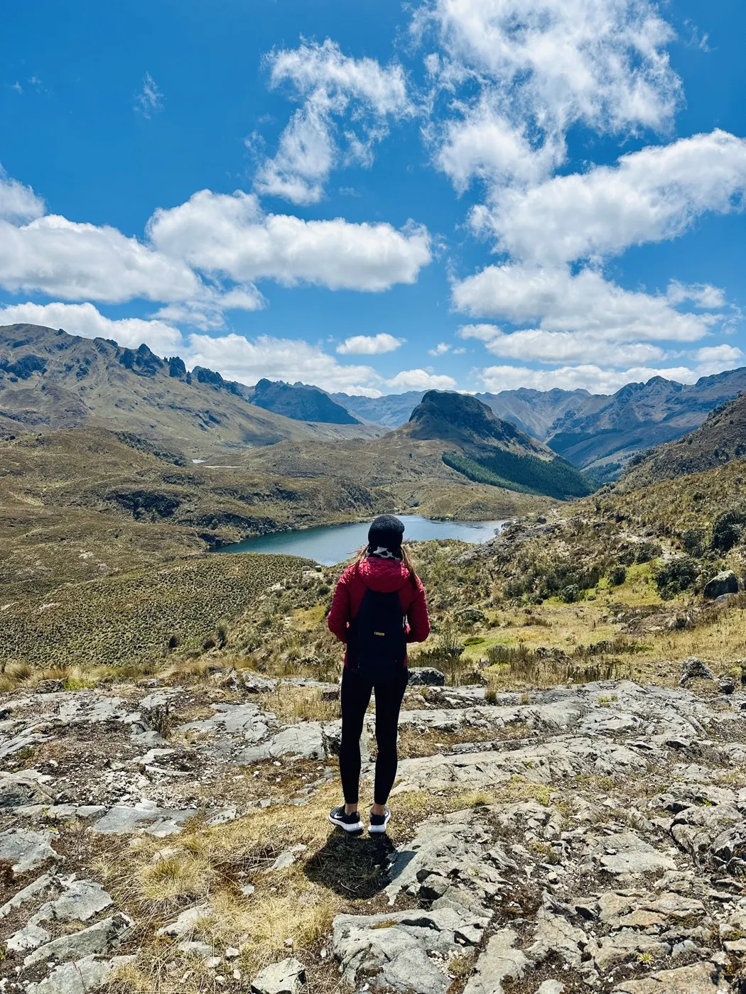 A lone hiker standing on a rocky outcrop above a high mountain lake ringed by peaks