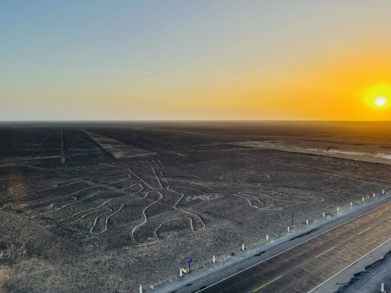 The sun setting over a vast desert plain crossed by ancient ground drawings, seen from a roadside viewing tower
