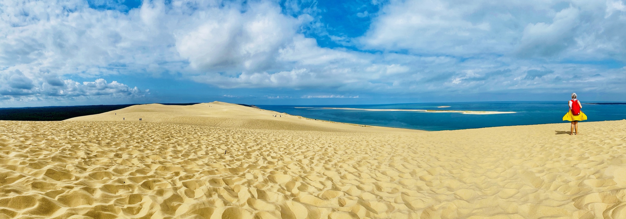 A traveler with a red backpack standing on a vast sand dune overlooking the sea
