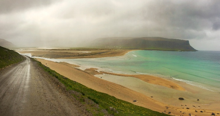 A lone gravel road tracing a misty Icelandic coastline, with turquoise shallows and a distant headland