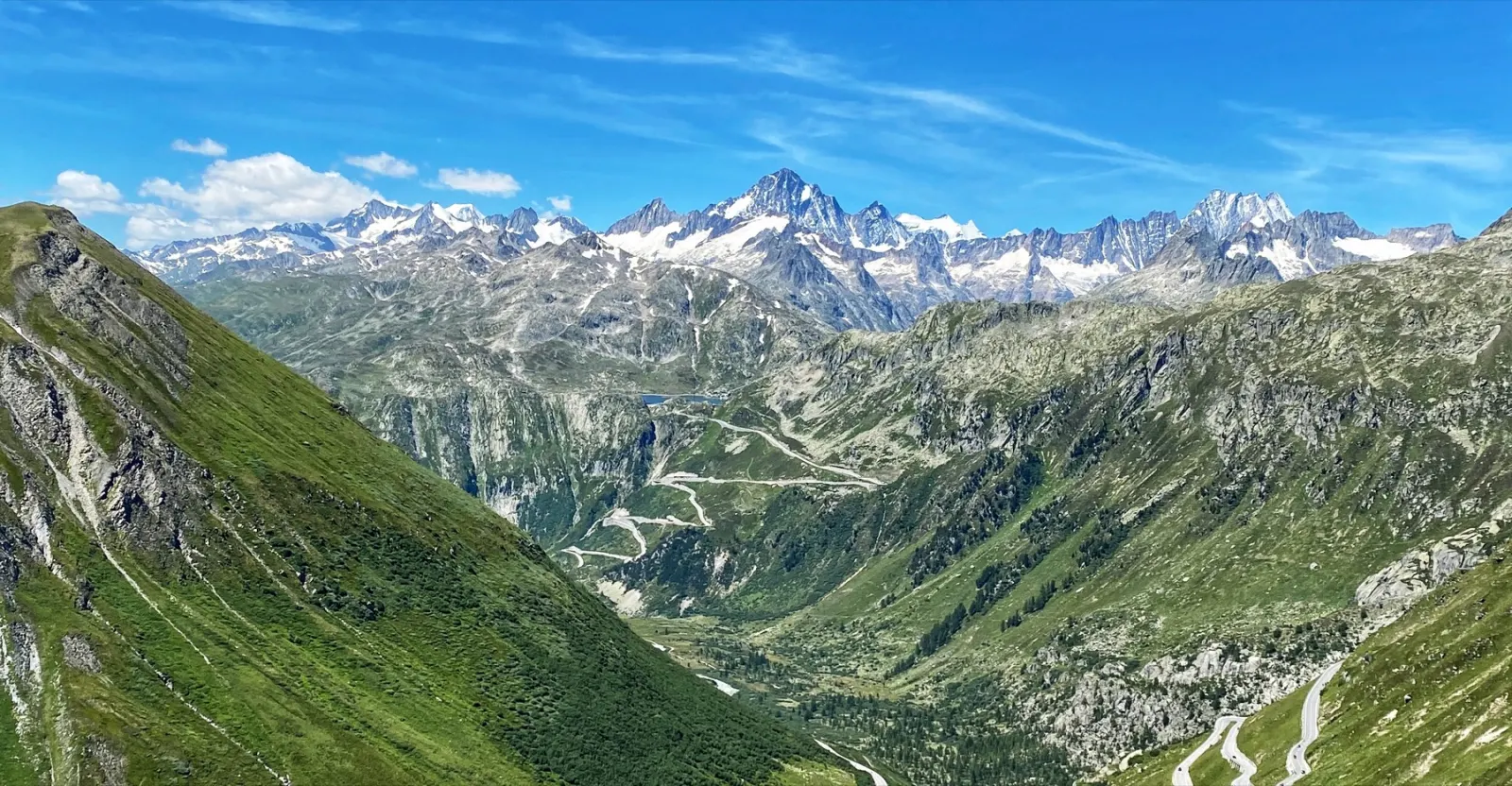A panorama of the Swiss Alps with snowy peaks rising above green slopes and hairpin pass roads winding through the valley below