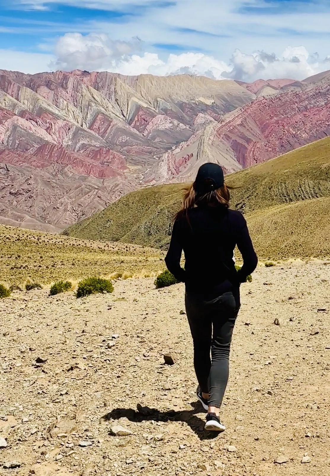 A lone walker heading across a desert ridge toward a wall of pink and ochre striped mountains